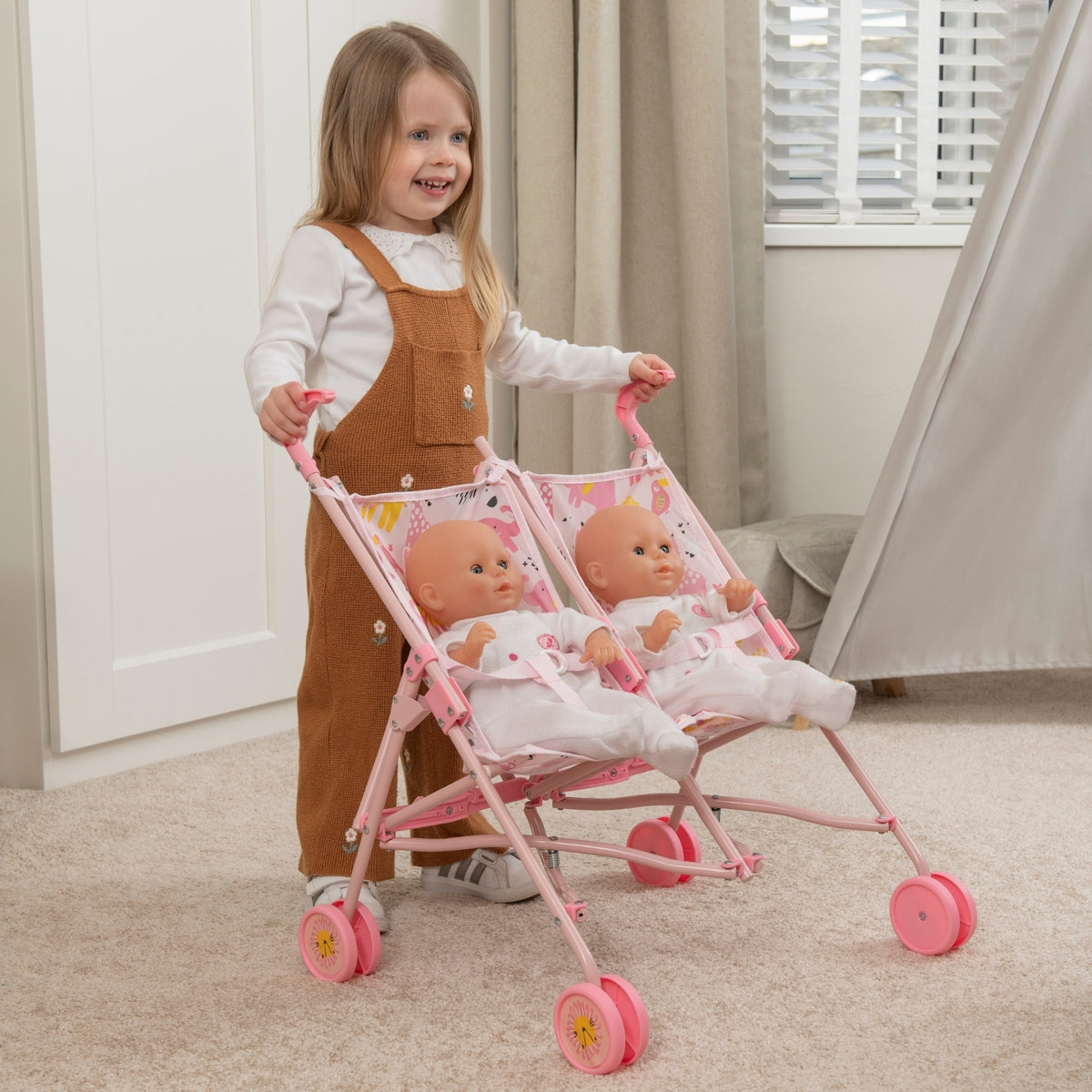 Child pushing a pink doll stroller with two dolls inside in a room.
