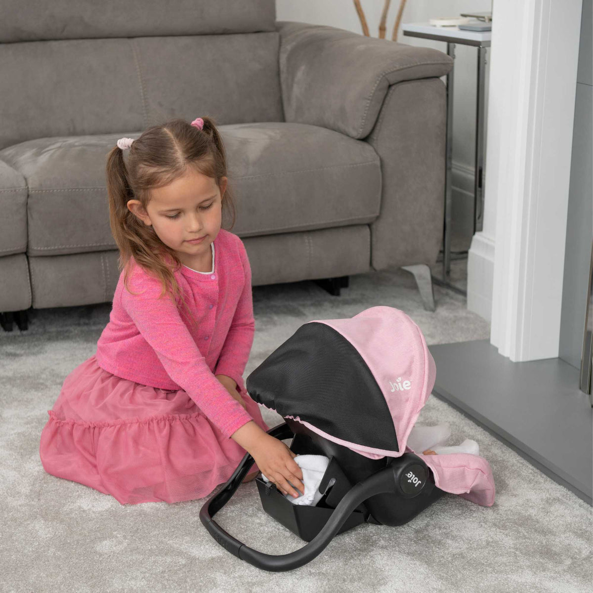 Joie I-Gemm Dolls Car Seat in pink and black, featuring a realistic design with secure harness, padded interior, and carry handle, perfect for children to safely transport their dolls during playtime